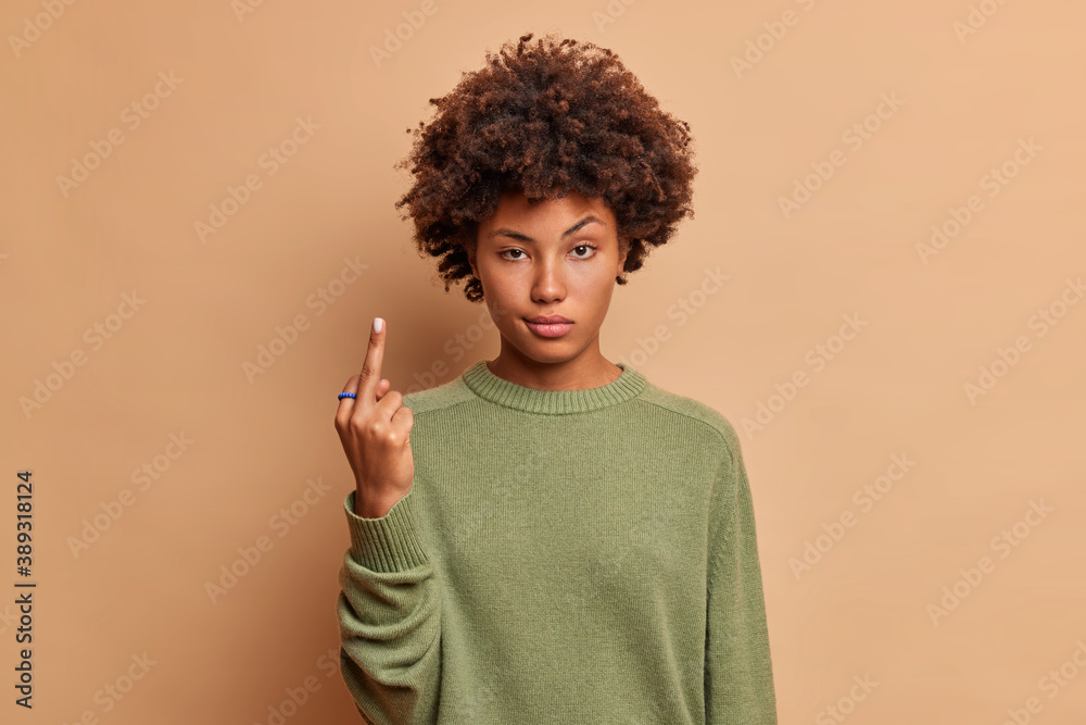 Serious dark skinned woman shows fuck you sign looks with poker face being vulgar and has quarrel with someone dressed in casual sweater isolated over beige wall. Female demonstrates middle finger Serious dark skinned woman shows fuck you sign looks with poker face being vulgar and has quarrel with someone dressed in casual sweater isolated over beige wall. Female demonstrates middle finger