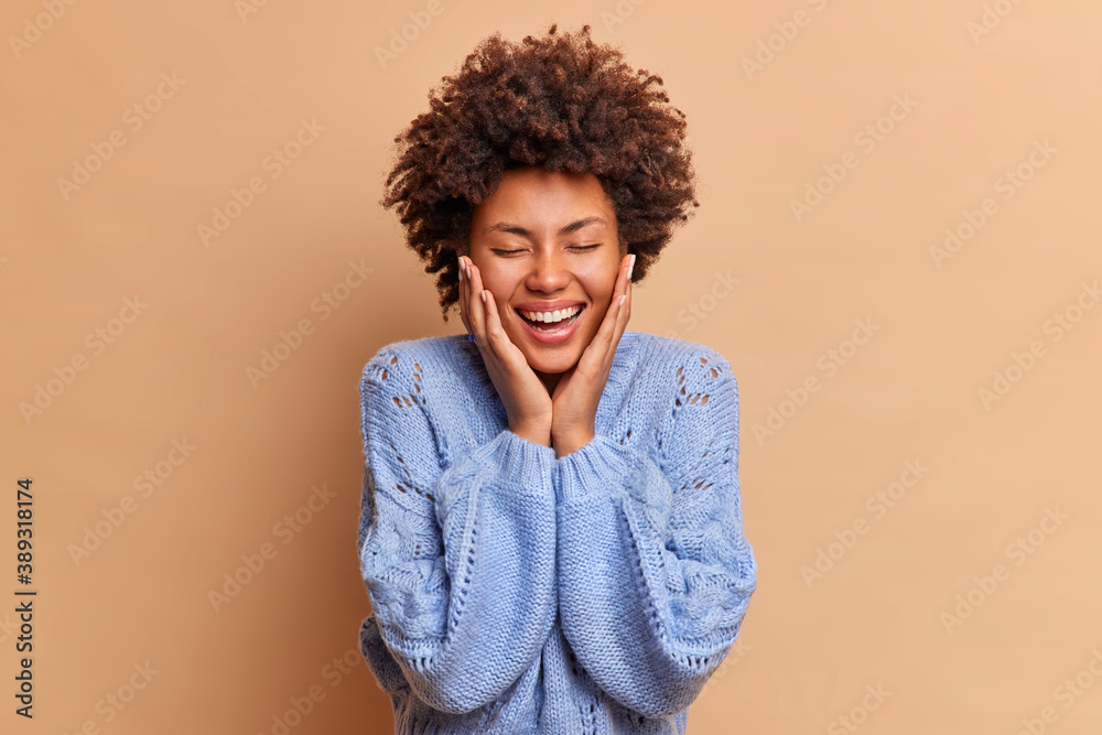 Human sincere feelings and emotions concept. Positive Afro American woman keeps hands on cheeks smiles broadly shows white perfect teeth feels joy or satisfaction has hilarious conversation. Human sincere feelings and emotions concept. Positive Afro American woman keeps hands on cheeks smiles broadly shows white perfect teeth feels joy or satisfaction has hilarious conversation.