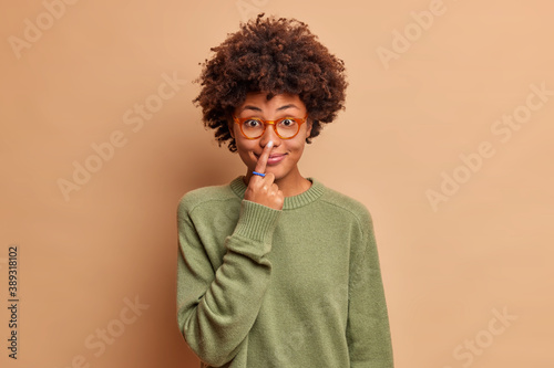 Funny curly haired young woman tries to touch nose smiles happily has fun and poses in casual green wear against brown background. Pretty teenage girl in spectacles with Afro hair points to nose.