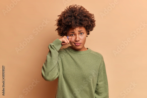 Gloomy dissatisfied curly haired woman wipes teardrops under eye expresses sadness and depression feels unhappy wears casual jumper isolated over brown studio wall. Negative emotions concept