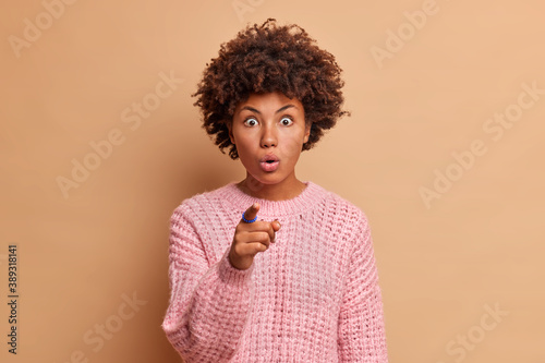 Shocked impressed woman with curly hair looks with great wonder points directly at camera sees something unbelievable poses speechless indoor dressed in knitwear isolated over beige background