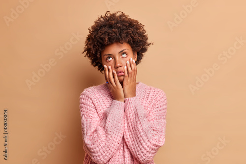 Upset bored Afro American woman keeps hands on cheeks suffers from boredom doesnt know what to do has dull expression stays at home alone dressed in casual sweater isolated over beige background © Wayhome Studio