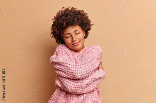 Fototapeta Pretty dark skinned curly woman embraces herself and closes eyes feels comfort in soft warm knitted sweater enjoys home tenderness tilts head poses against beige background