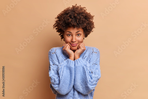 Good looking African American woman with natural curly hair keeps hands under chin dressed in blue jumper looks directly at camera stands confident and beautiful stands against brown background