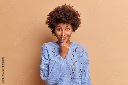 Pretty dark skinned African American woman touches nose with index finger smiles cheerful has funny look at camera dressed in casual blue jumper isolated over brown background. Look how I can © wayhome.studio 