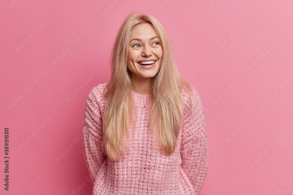 Joyful blonde woman smiles sincerely looks away with glad expression recalls something funny happened dressed in knitted jumper isolated over pink background laughs from amusement has thoughtful look Joyful blonde woman smiles sincerely looks away with glad expression recalls something funny happened dressed in knitted jumper isolated over pink background laughs from amusement has thoughtful look