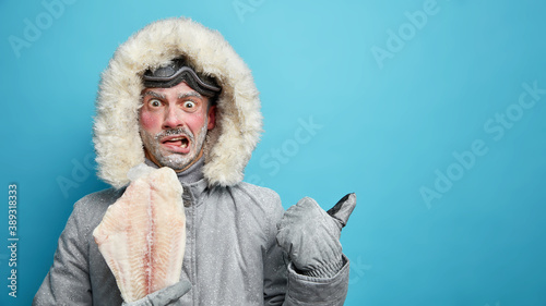 Puzzled angry man with frozen face during winter fishing dressed in grey jacket with hood and gloves points away on free space against blue background shows way to mountain lake. Northern lifestyle