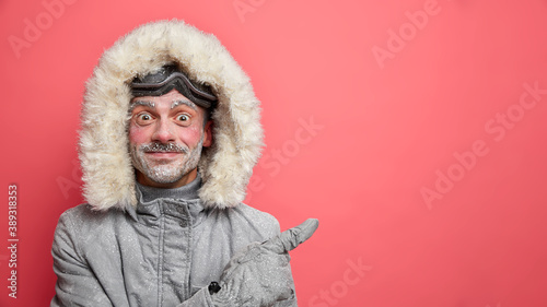 Happy bearded snowboarded covered with snow has active rest in mountains points at copy space against vivid rosy background dressed in warm outerwear. Be ready for frosty days. Winter is coming