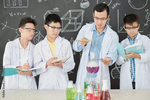 Science teacher demonstrating chemical reaction to students, who are taking notes when he is pouring reagent in laboratory flask