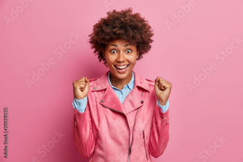 Overjoyed excited young woman with Afro hair clenches fists as sign of victory enjoys moment of success anticipates for something intriguing celebrates great result poses against rosy background