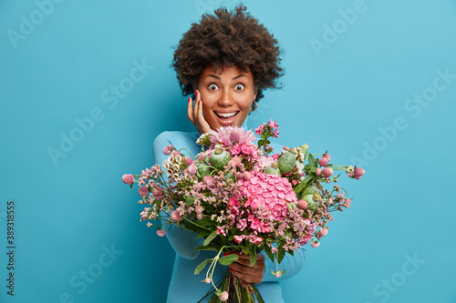 Cute cheerful woman touches face gently holds big bunch of bouquet going to congratulate best friend with special occasion poses against blue background. My best 8 march ever. Beautiful spring girl