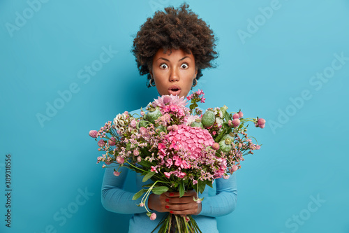 Young shocked curly haired woman receives festive bouquet in honor of womens day stands speechless indoor against blue background. Stunned florist prepares bunch of flowers for special occasion.