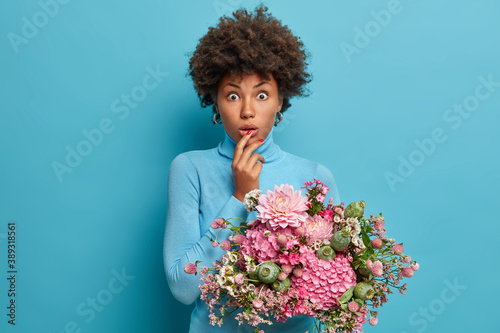 Photo of surprised dark skinned woman gets flowers from stranger dressed in casual wear isolated over blue background. Afro American lady celebrates birthday or spring holiday. Florist with bouquet