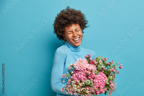 Beautiful overjoyed Afro American woman in casual turtleneck receives bouquet of flowers as gift expresses positive emotions isolated over blue background celebrates birthday or special event.