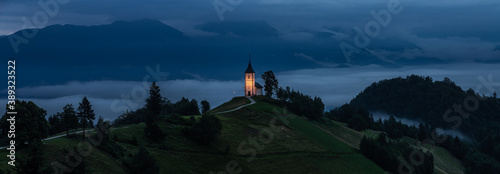 Jamnik, Slovenia - Panoramic view of blue hour at Jamnik with illuminated St. Primoz hilltop church on a foggy summer dawn. Julian Alps at background