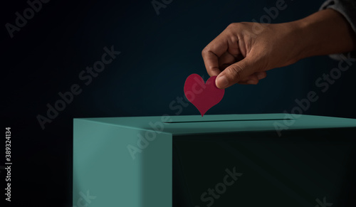 Donation Concept. Hand putting a Red Heart paper into a Donate Box. Metaphor photo. Dark tone photo