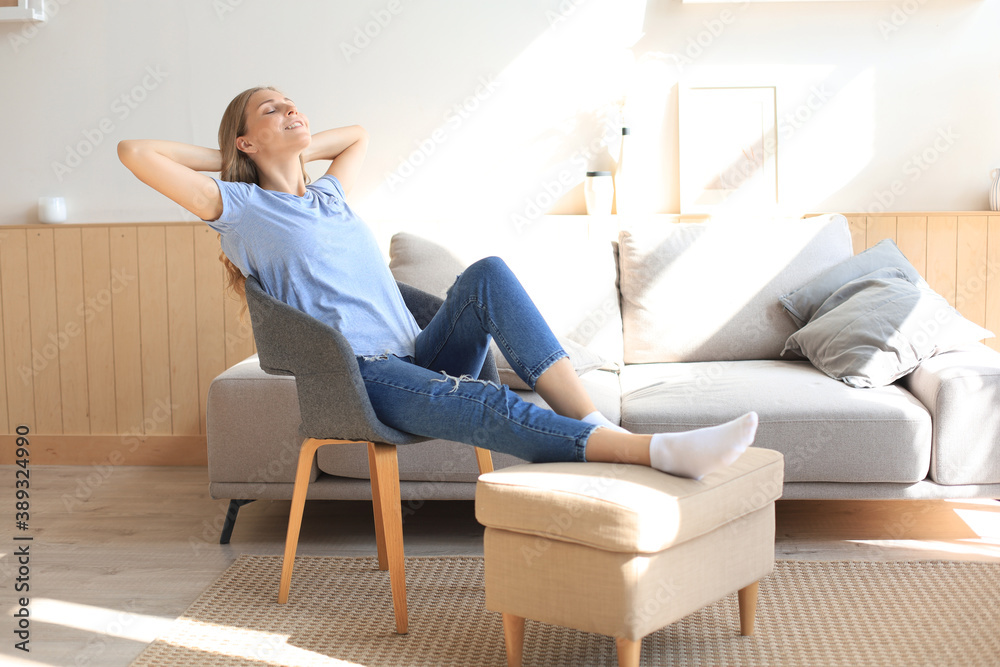 Young woman at home sitting on modern chair in front of window, relaxing in her living room.