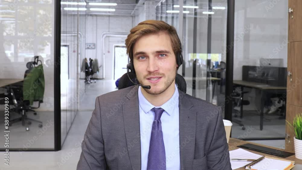 Smiling male call-center operator with headphones sitting at modern office, consulting online information, assistance to the client, looking at camera.
