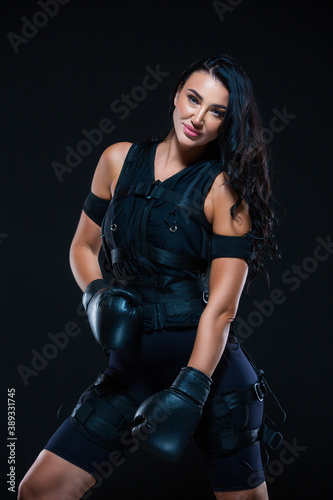 A young athletic woman in an EMS suit with Boxing gloves on an isolated black background. EMS training. Electro muscular stimulation machine.