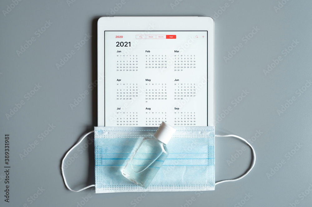 a tablet with an open calendar for 2021 year and protective medical mask and hand sanitizer on a gray background. covid-19 coronavirus protection concept in 2021 a tablet with an open calendar for 2021 year and protective medical mask and hand sanitizer on a gray background. covid-19 coronavirus protection concept in 2021