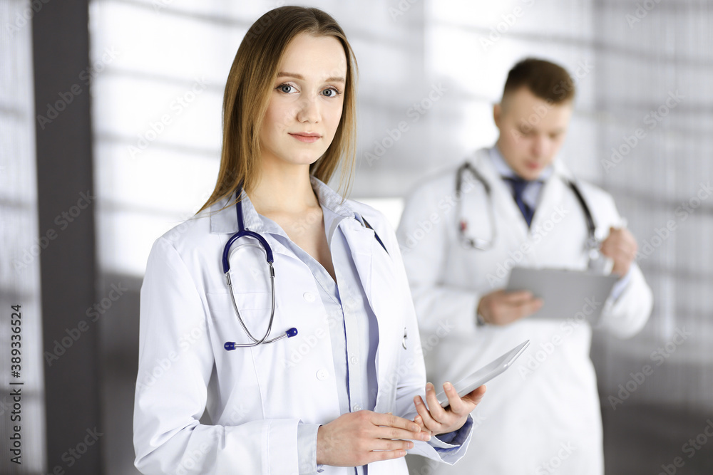 Professional woman doctor with a stethoscope is using a computer tablet, while she is standing together with her colleague in a clinic. Young doctors at work in a hospital. Medicine and healthcare Professional woman doctor with a stethoscope is using a computer tablet, while she is standing together with her colleague in a clinic. Young doctors at work in a hospital. Medicine and healthcare