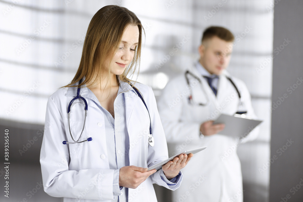 Professional woman doctor with a stethoscope is using a computer tablet, while she is standing together with her colleague in a clinic. Young doctors at work in a hospital. Medicine and healthcare Professional woman doctor with a stethoscope is using a computer tablet, while she is standing together with her colleague in a clinic. Young doctors at work in a hospital. Medicine and healthcare