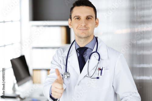 Male doctor standing with thumbs up sign in clinic near his working place. Perfect medical service in hospital. Medicine and healthcare concept