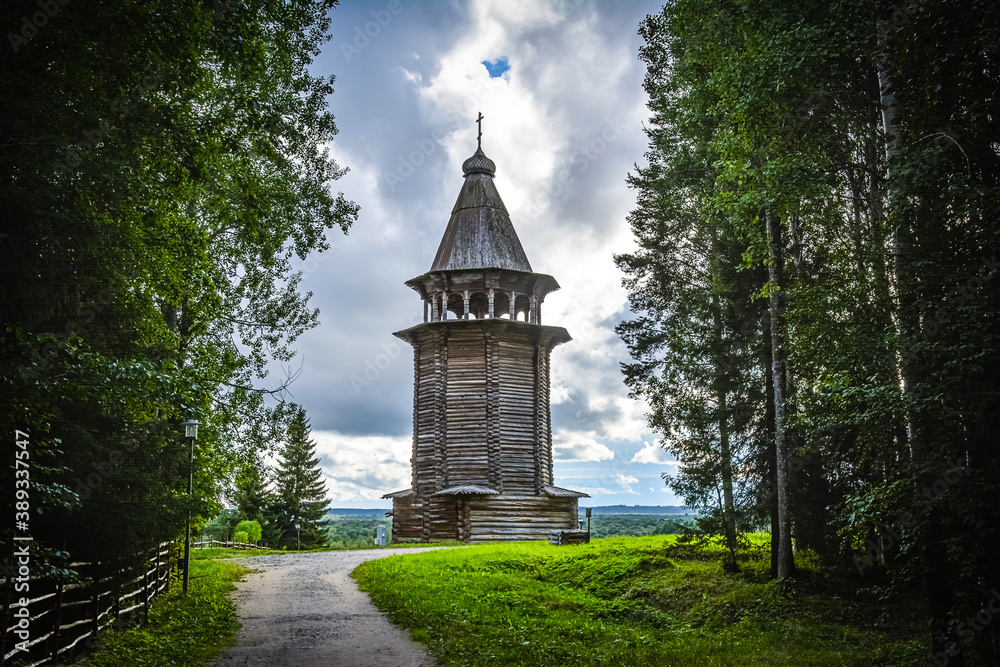 view from the dark forest of the wooden bell tower against the sky with clouds view from the dark forest of the wooden bell tower against the sky with clouds