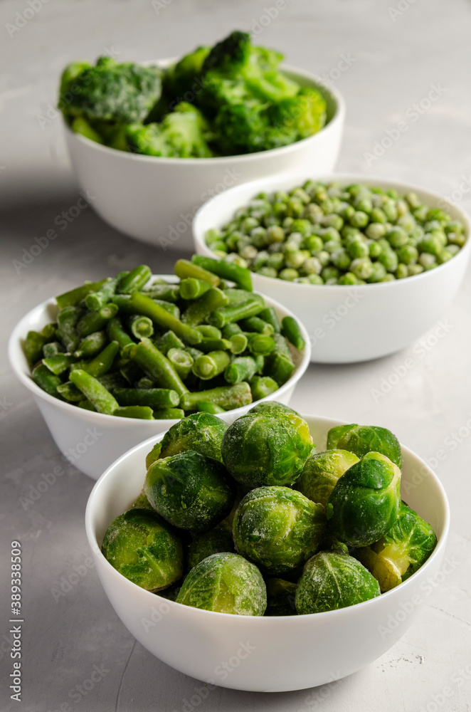 A mix of frozen green vegetables: beans peas broccoli Brussels sprouts in bowls on a grey back A mix of frozen green vegetables: beans peas broccoli Brussels sprouts in bowls on a grey back