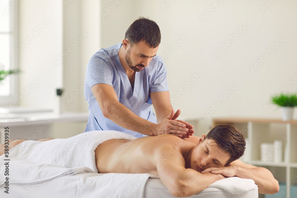 Serious masseur doing relaxing back massage for young man using Tapotement technique - obrazy, fototapety, plakaty 