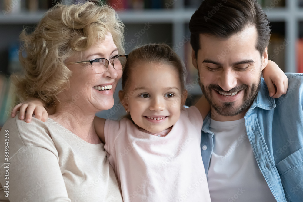 Happy three generations bonding family enjoying sweet tender moment at home. Head shot smiling adorable small child girl cuddling affectionate 60s older granny and caring young father indoors.