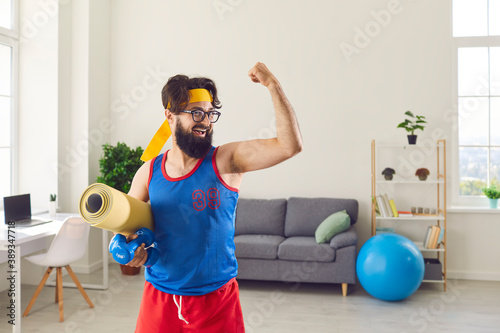 Fototapeta Naklejka Na Ścianę i Meble -  Funny athlete holding gym mat and dumbbells and showing his weak biceps after sport workout at home