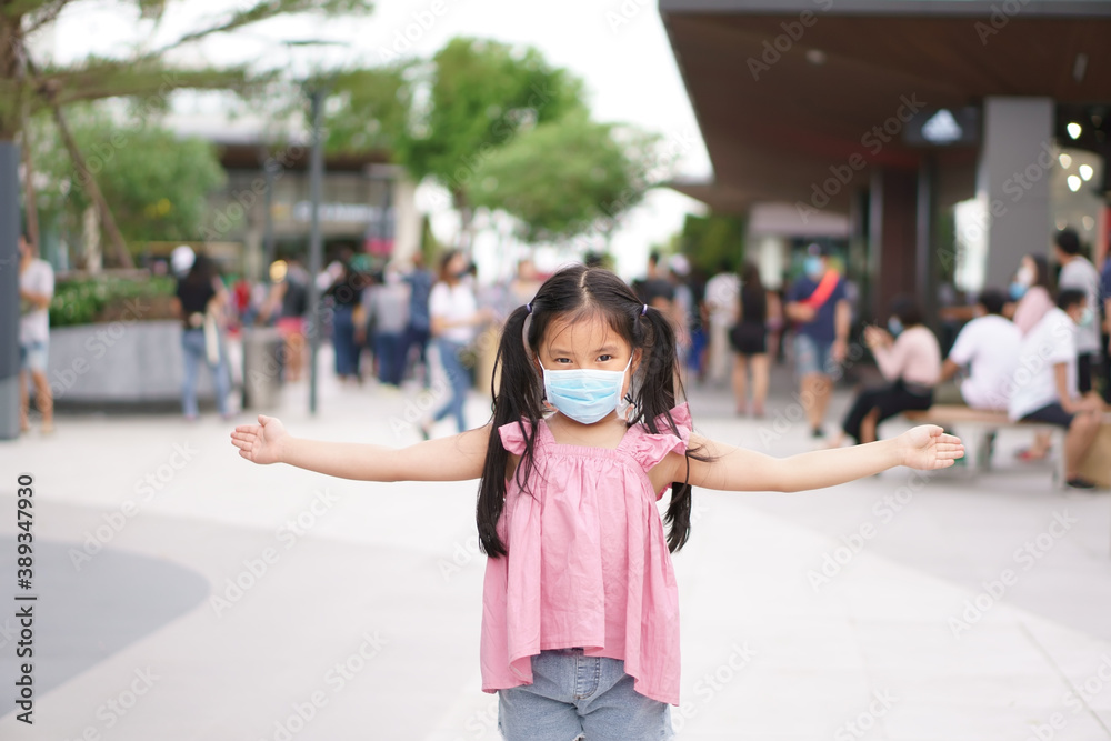 Asian child or kid girl wearing blue face mask and open arm to present or stop no entry on lot of people for social distancing on shopping mall or department store on holiday in coronavirus covid-19