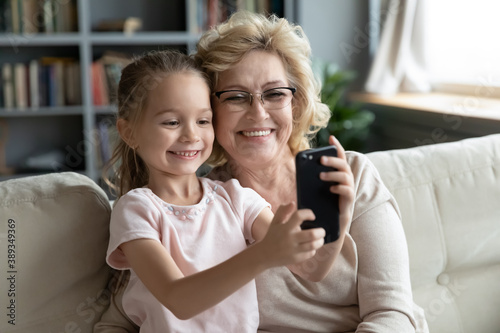 Happy middle aged 60s grandmother in glasses posing for selfie on smartphone with adorable small granddaughter at home. Little cute kid girl recording funny mobile video with granny or making call.