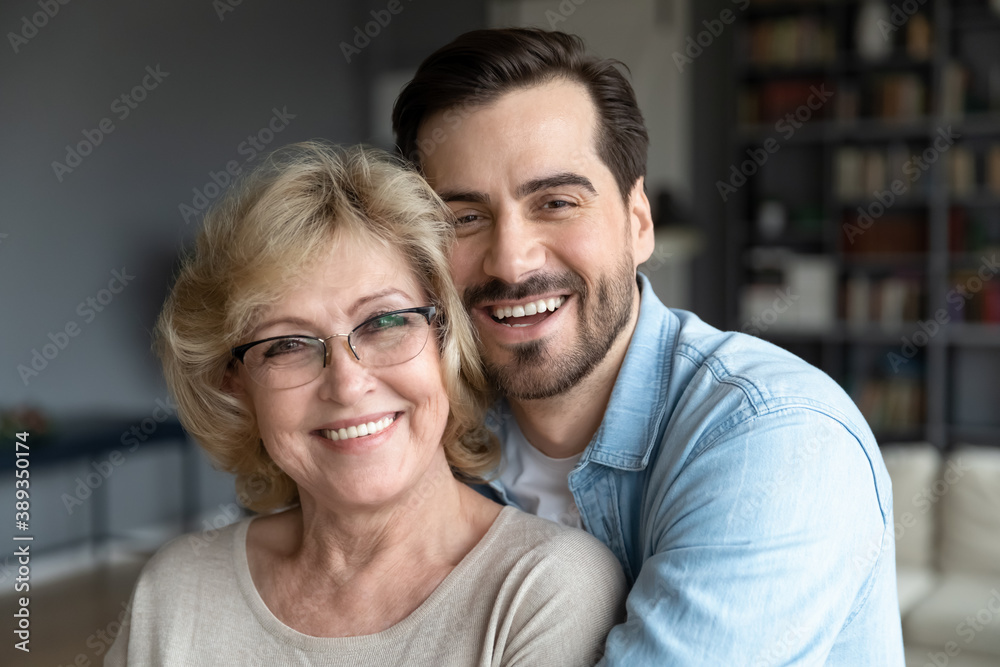 Head shot close up portrait of loving young bearded man cuddling smiling beautiful middle aged mature mother on eyeglasses. Loving different generations family looking at camera, enjoying weekend.