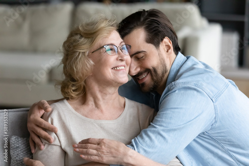 Young caring son cuddling happy elderly senior retired mother in eyeglasses, feeling thankful at home. Smiling 60s mature woman enjoying sincere trustful relations moment with grown up child at home.