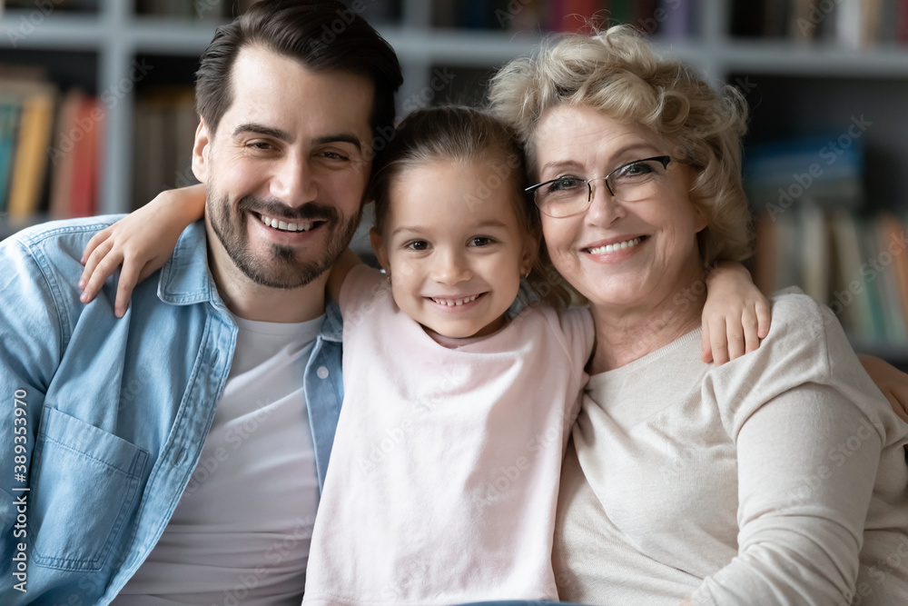 Head shot portrait of bonding smiling intergenerational family of three. Happy little preschool child girl embracing affectionate young father and elderly retired 60s grandmother, looking at camera.