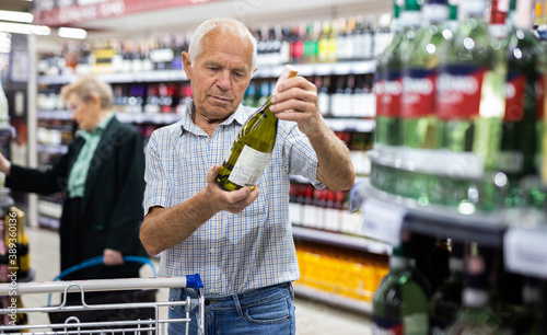 mature european man chooses bottle of white wine in alcohol section of supermarket