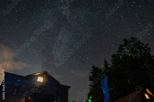 Lonely glowing window in the house. Silhouette of a house and a dried tree against the background of the constellations and the Milky Way. Dramatic night scenery with a starry sky.