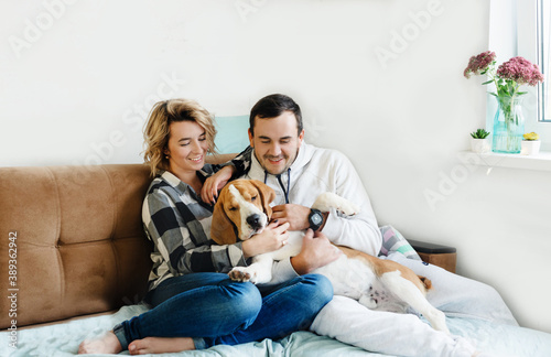 Happy young couple hugs two cute dogs sitting on the sofa. The youth way of life, the concept of home and animals. © Tatsiana