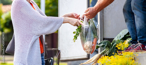 Young anonymous woman buying fresh vegetables, groceries from the mobile greengrocer in his truck. Passing the plastic bag full of greens from hand to hand. Healthy eating, simple farm trade concept photo