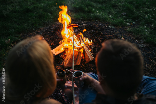 Sausage roasting on fire. Couple cooking together. Bonfire cooking in forest. Campfire picnic outdoor. Heating up food on wooden stick. Natural eco barbecue meat smoking. Yerba mate drink in nature.