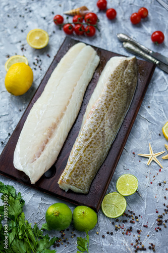 fillet of white fish two pieces on a wooden board on a gray background with knives lemons and tomatoes 