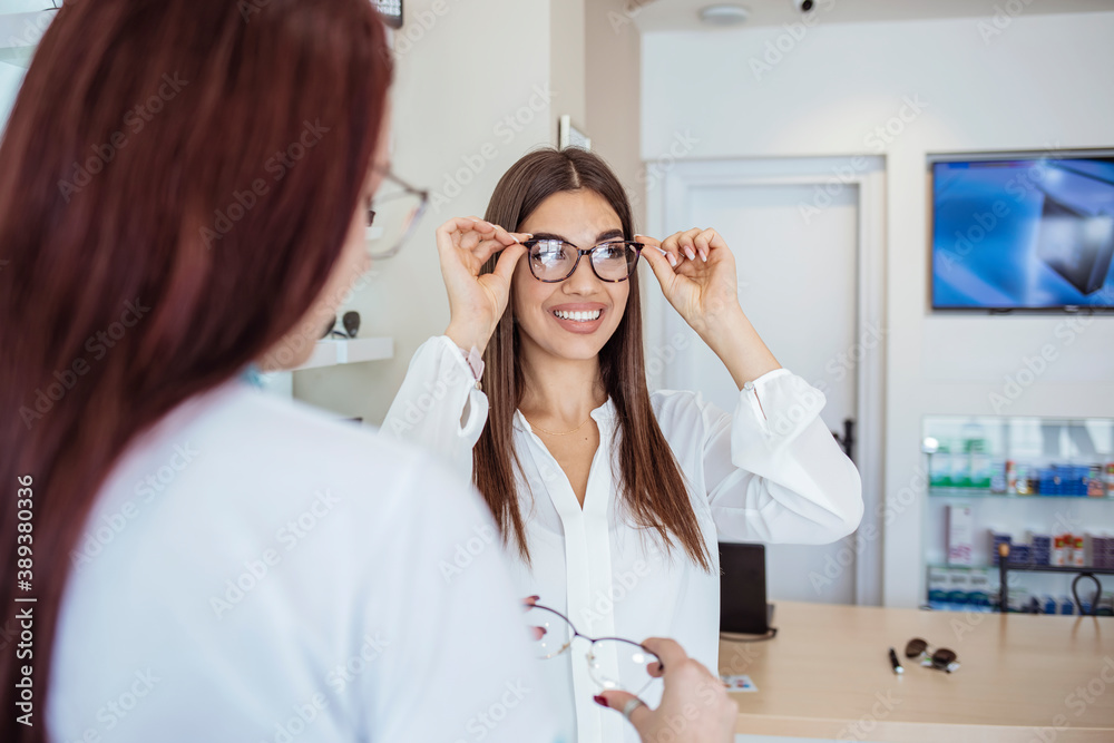 Young smiling woman is trying on eyeglasses in optical shop with the assistance of optometrist. Woman trying on eyeglasses in optical shop. Joyful happy woman trying some eyeglasses Young smiling woman is trying on eyeglasses in optical shop with the assistance of optometrist. Woman trying on eyeglasses in optical shop. Joyful happy woman trying some eyeglasses
