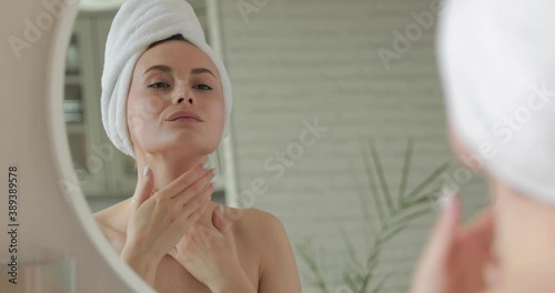 Pleasant young lady wrapped in bath towel smoothing her skin with face cream while looking at mirror at bright bathroom. Concept of morning routine at home.