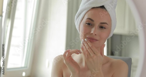 Portrait of happy young woman with towel on head doing facial massage while standing in modern bathroom. Beautiful female touching skin gently with fingers.