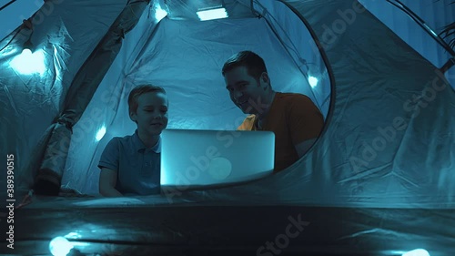 Father and son playing in tent with Christmas decorations. Family watching movies on laptop, having fun in evening holidays time at home.