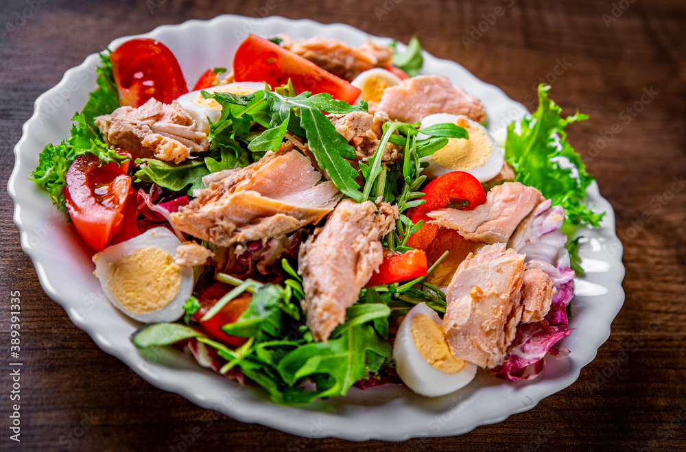 Fresh salad with fish, arugula, eggs,red pepper, lettuce, fresh sald leaves and tomato on a white plate on wooden table background