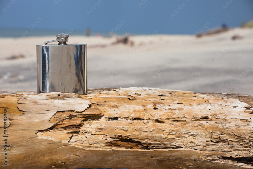 Hip flask on a wooden beam lying on the beach. - obrazy, fototapety, plakaty 