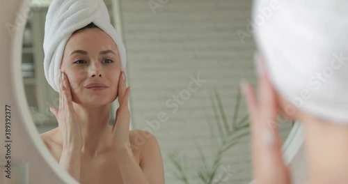 Happy lady in white towel standing in bathroom and applying cream on face. Young woman with bare shoulders using korean cosmetics for skin care.